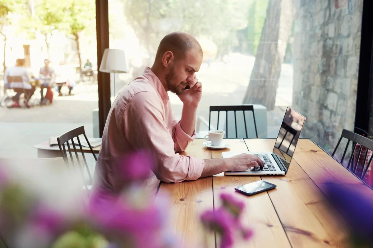 Man in a pink shirt sitting at a wooden table using a laptop and talking on a phone in a bright cafe.