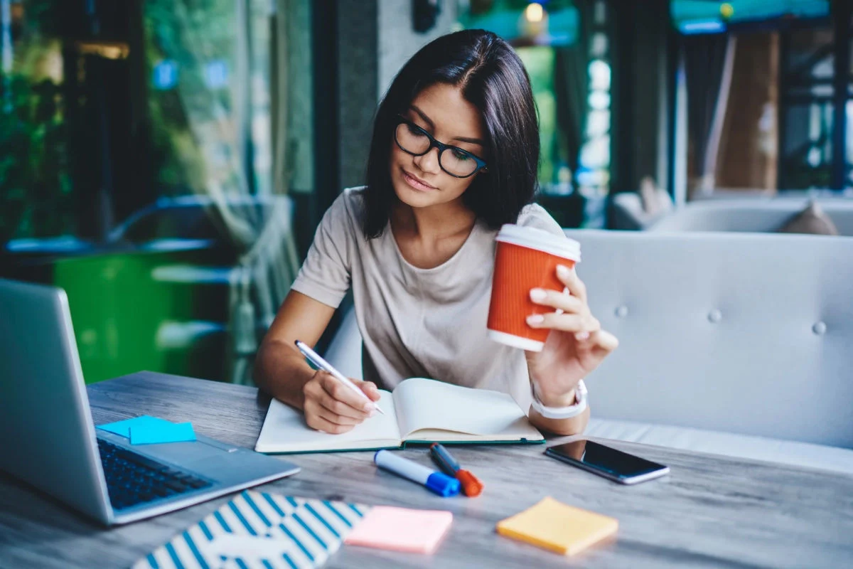Young woman with glasses sitting at a table in a cafe, writing in an open notebook with a pen while holding a red disposable coffee cup, with a laptop, phone, markers, and sticky notes on the table.