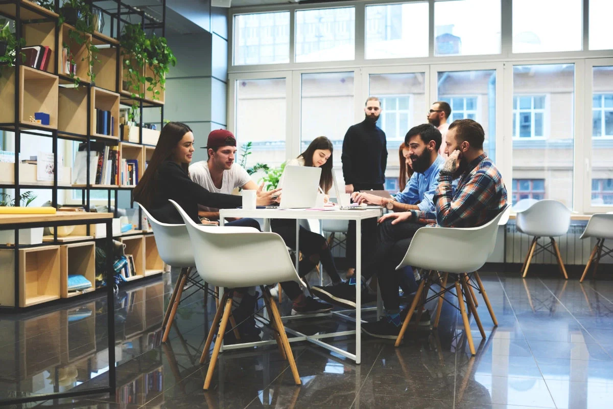 A group of young adults sitting around a table in a modern office space, working on laptops and engaging in discussion.